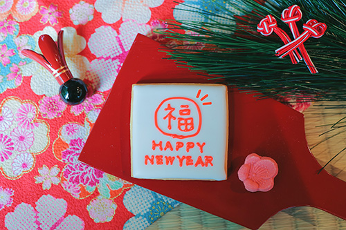 フリー写真画像『【年賀状画像】羽子板に乗った『HAPPY NEWYEAR』のアイシングクッキー（赤和柄背景）』[ID:10864]